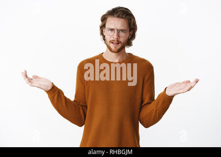 Vita-up shot di dispiaciuto e deluso bella giovane maschio in bicchieri e maglione marrone accigliata con le mani alzate lateralmente e spallamento Foto Stock