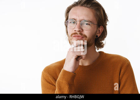 Studio shot di esitanti e sicuri di buona cercando giovani redhead ragazzo con naso forato e orecchini smirking dubbia tenendo la mano sulla barba rendendo Foto Stock