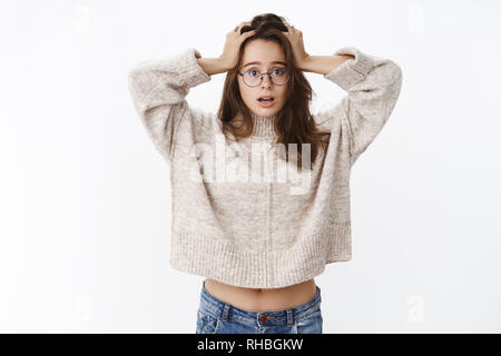 Oh mio, cosa fare. Sciocca e clueless bella ragazza confusa in bicchieri e maglione tenendo le mani sul capo, bocca aperta dalla sorpresa, inconsapevoli come Foto Stock