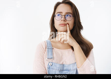 Ragazza pensando cosa ordinare per cena, tenendo il dito sul labbro guardando pensieroso ad angolo superiore sinistro come rendere la decisione strizzare gli occhi concentrata in piedi Foto Stock