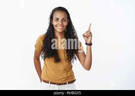 Affascinante timido e timido afro-donna americana con lunghi capelli ricci in senape t-shirt sollevando il dito indice rivolto verso l'alto come azienda mano dietro insicuro Foto Stock