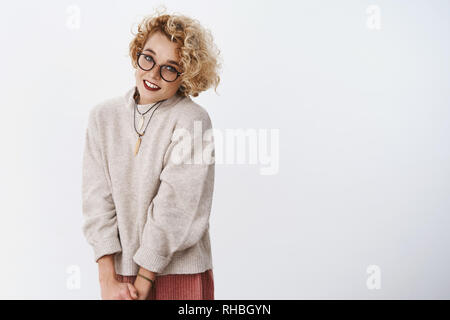 Portarit di carino e timido flirty ragazza bionda in bicchieri e maglione rendendo graziosa sguardi a telecamera sensualmente sorridente e femminile come in posa su bianco Foto Stock