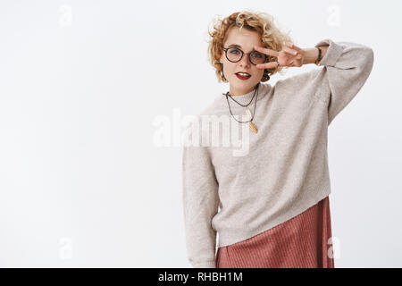 Ritratto di fresco ed elegante tanga giovane ragazza in felpa trendy e i bicchieri che mostra il gesto di pace o discoteca segno over eye e guardando la telecamera di profondità Foto Stock