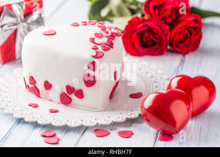 Il marzapane torta bianca a forma di cuore con cuori rossi. Come decorazione bouquet di rose rosse un regalo dal nastro. Il matrimonio o il giorno di san valentino Foto Stock