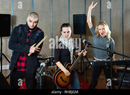 Tre positivi felice compagni di band che pongono insieme con gli strumenti musicali in sala prove Foto Stock