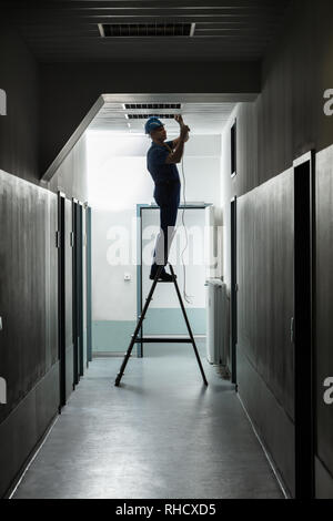 Silhouette di un elettricista maschio sulla scaletta Installazione di luce in corridoio Foto Stock