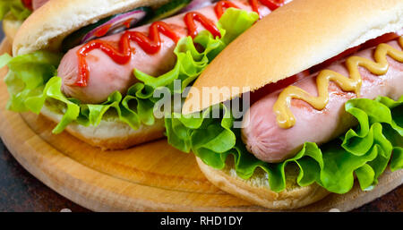 Deliziosi hot dog con salsiccia grill, ketchup, senape, lattuga, pomodori su un vassoio in legno Close up Foto Stock