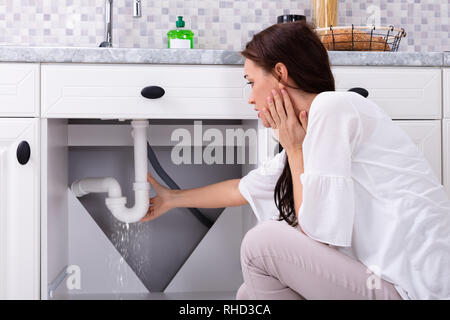 Vista posteriore di una donna che cerca di fermare la perdita di acqua dal tubo di lavello in cucina Foto Stock
