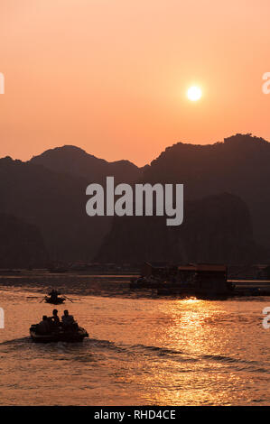 La gente del luogo in piccole casette di legno barca da pesca e villaggio di pescatori stagliano tra pietre calcaree carsiche come il sole tramonta sulla baia di Ha Long, Vietnam Foto Stock