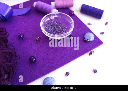 Un set di materiali per il ricamo in colore viola su uno sfondo bianco. Perle, filettature, un nastro di raso e lana per maglieria. Foto Stock