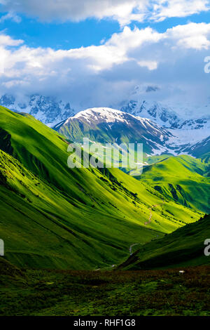 Verdi campi e colline che portano a montagne coperte di neve. Nuvole bianche sono visibili dietro gli illuminati di montagna. Estate in Georgia caucasica. Verti Foto Stock