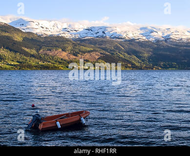 Red motoscafo sulle sponde di un lago. Sull'altro lato case e snowy mountain range sotto i raggi del sole di sera. L'acqua è blu, ondulate. Gabbiano seduta sul Foto Stock