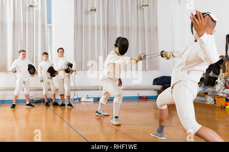 Giovani schermitori guardando la scherma duello con lamine di due allenatori professionisti in palestra Foto Stock