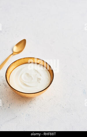 Ciotola di fresco lo yogurt greco e il cucchiaio d'oro bianco su sfondo astratto. Una sana prima colazione. Golden utensili. Vista dall'alto. Copia spazio per il testo. Foto Stock