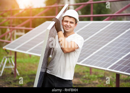 Sorridente giovane lavoratore nel casco protettivo che trasportano big shiny solar photo voltaic sul pannello esterno piattaforma metallica sullo sfondo estiva soleggiata giornata calda. Fonti ecologiche di energia verde concetto. Foto Stock