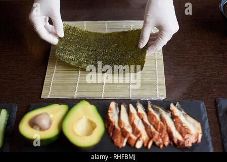 Vista da vicino il processo di preparazione del sushi di laminazione. Lo chef ha le mani tenendo foglio di nori. Avocado e anguilla su nero piastre di pietra Foto Stock