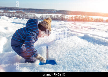 Un piccolo ragazzo bambino gioca nella neve in inverno. Spazio libero per il testo. In tuta blu e cappello. Guarda la pala in un cumulo di neve. Foto Stock