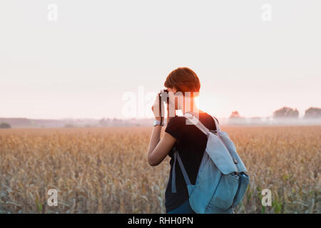 Retroilluminato con la figura di una donna di scattare una foto in brillante tramonto. Persona di sesso femminile con zaino in piedi di sera la luce del sole in un campo e utilizzando analo vintage Foto Stock