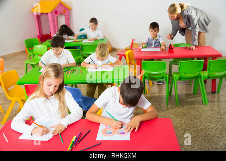 Schoolkids funzionante a scrivanie in aula e insegnante che li aiuta Foto Stock