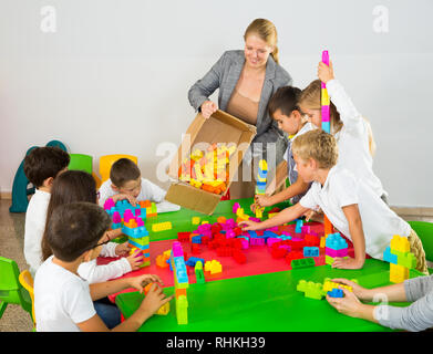Gruppo di schoolkids giocando con mattoni giocattolo in cerchio con il loro allegro insegnante femmina Foto Stock