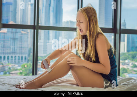 Giovane donna sitin sul letto di casa e facendo l'epilazione con epilatore sulle gambe ed è nel dolore. Sullo sfondo di una finestra che si affaccia sulla grande città Foto Stock