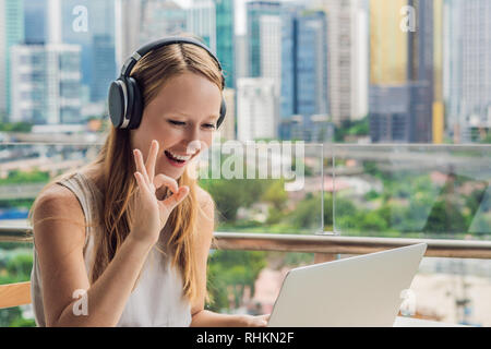 Giovane donna insegna una lingua straniera o impara una lingua straniera in Internet sul suo balcone contro il fondale di una grande città. Lingua on line Foto Stock