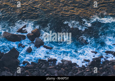 Deep Blue onde dell oceano Atlantico colpendo il basalto scuro shore con luce solare d'oro riflettente onde off al tramonto. Capo Roca Foto Stock