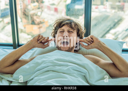 Giovane uomo disteso su un letto coperto le orecchie a causa del rumore. Nella finestra dopo il letto è possibile vedere la costruzione di una nuova casa Foto Stock