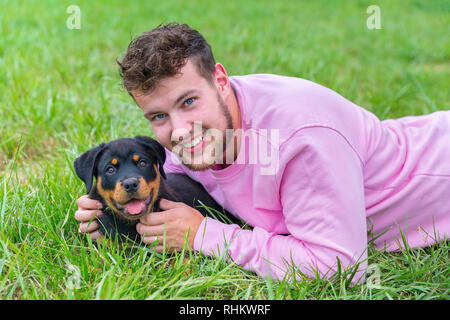 Giovane maschio caucasico proprietario del cane tiene amorevolmente rottweiler cucciolo in prato naturale Foto Stock