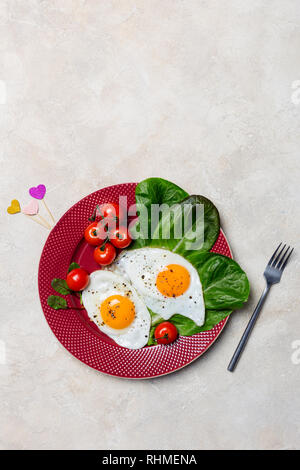 Colazione romantica con due uova fritte, pomodori ciliegini, insalata romano a targhetta rossa, a forma di cuore ad stick, forcella vicino a sfondo chiaro. Libera verticale s Foto Stock