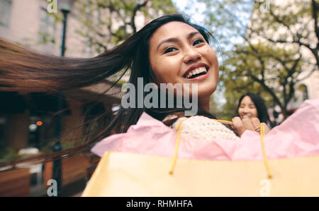 Vista posteriore di un sorridente donna asiatica tornando portando una borsa da shopping. Donna allegra che porta un sacco di shopping all'aperto a piedi con i suoi capelli battenti Foto Stock