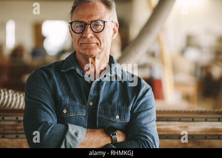 Ritratto di successo uomo senior in occhiali in piedi nel suo laboratorio di falegnameria. Falegnameria orgoglioso proprietario di officina in piedi con le braccia incrociate. Foto Stock