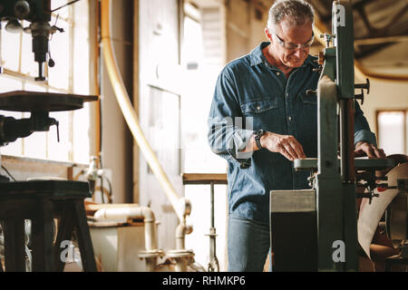 Maschio maturo falegname lavorando su una sega a nastro macchina nel laboratorio di falegnameria. Senior uomo taglio del legno sulla macchina. Foto Stock