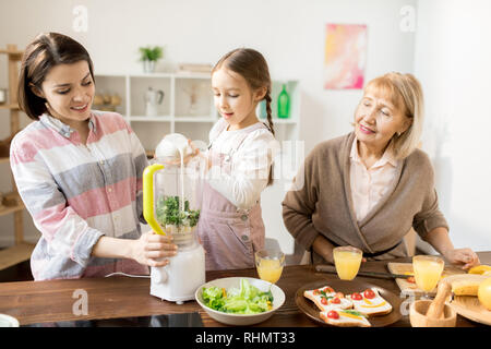 Ragazza Adorale versando il latte fresco nel frullatore elettrico mentre aiuta la sua mamma con rendendo i frullati Foto Stock