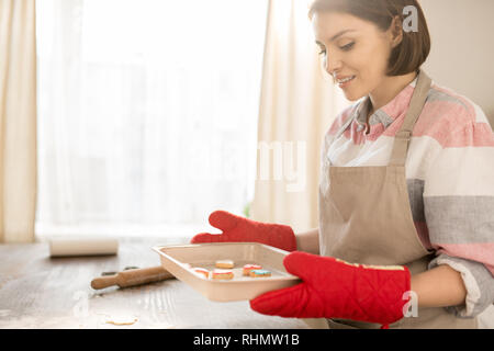 Piuttosto giovane massaia in grembiule e guanti di protezione tenendo il vassoio a caldo con biscotti fatti in casa prese al di fuori del forno Foto Stock