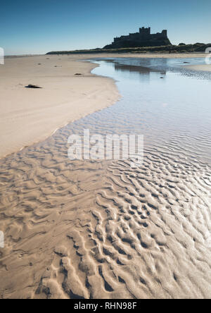 Il castello di Bamburgh riflessa nelle acque poco profonde sulla spiaggia con sabbia ondulazioni in primo piano. Foto Stock