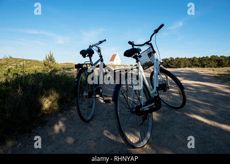 Due biciclette parcheggiate sulla strada; la Chiesa Sand-Covered in background contro il cielo blu a Skagen, nello Jutland, Danimarca. Foto Stock