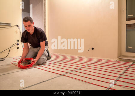 Elettricista installazione di riscaldamento elettrico rosso il filo del cavo sul pavimento di cemento in camera incompiuto. Ristrutturazione e costruzione, confortevole casa calda concep Foto Stock