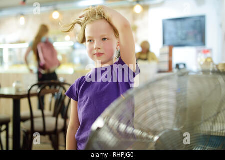 Carino bambina in piedi di fronte ad un ventilatore sul caldo giorno d'estate. Bambino godendo del vento fresco nella stagione estiva. Hot condizioni atmosferiche. Foto Stock