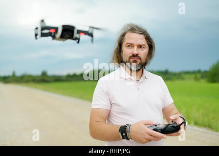 L'uomo la visione e la navigazione di un drone. Uomo Barbuto il funzionamento del drone dal telecomando. Drone passando davanti a un maschio sul giorno di estate. Foto Stock