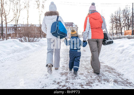 La mamma e la ragazza vede il ragazzo s figlio di 3-5 anni a mano a piedi giù per la strada. Vista posteriore. Inverno in città sullo sfondo di cumuli di neve. Tornando Foto Stock