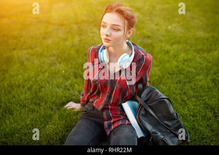 Bella giovane studente che indossa una camicia a scacchi le cuffie e uno zaino in appoggio sulla strada tra classi. Bella ragazza redhead Foto Stock