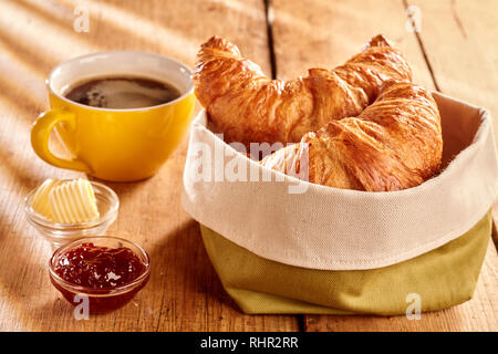 Pane appena sfornato sfaldabile croissant serviti nel sacco di tessuto con riccioli di burro, marmellata e caffè sulla tavola in legno rustico Foto Stock