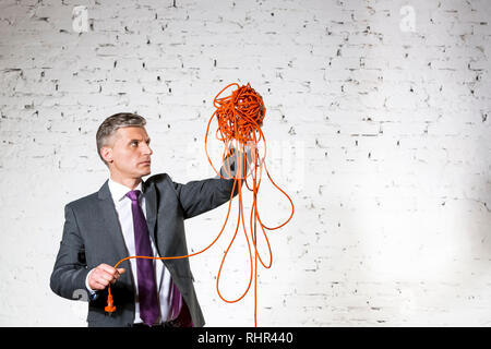 Fiducioso imprenditore maturo holding aggrovigliato cavo bianco contro un muro di mattoni Foto Stock