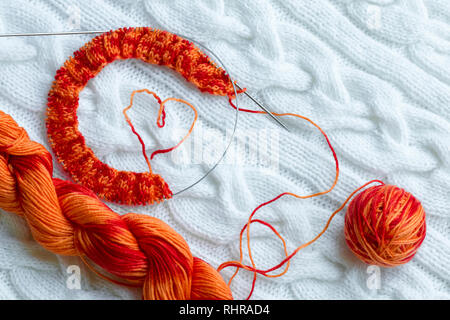 Progetto di lavorazione a maglia in corso. Un pezzo di maglia con una sfera e una matassa di colore rosso-arancio in sezione di tintura e di lavorazione a maglia sullo sfondo di un Foto Stock