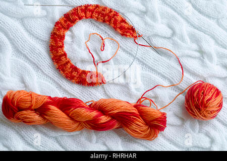 Progetto di lavorazione a maglia in corso. Un pezzo di maglia con una sfera e una matassa di colore rosso-arancio in sezione di tintura e di lavorazione a maglia sullo sfondo di un Foto Stock