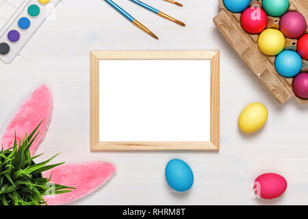 Un telaio, orecchie di coniglietto, un vaso di fiori artificiali con erba verde e colorati di uova in una scatola di cartone, vernici e pennelli. Concetto di pasqua. Spazio di copia Foto Stock