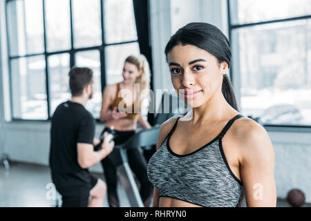 Bella African American sportive sorridente alla fotocamera, giovani amici formazione dietro in palestra Foto Stock