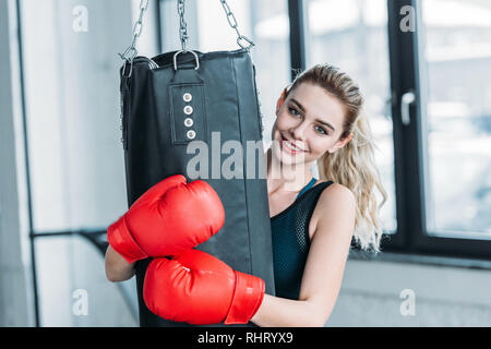 Felice ragazza sportiva in guantoni da pugilato abbracciando sacco da boxe e sorridente alla fotocamera in palestra Foto Stock