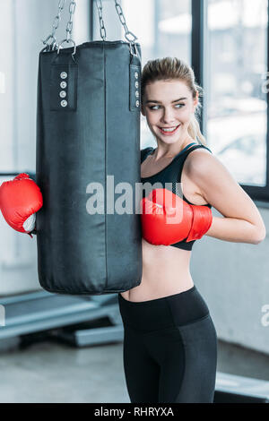 Bella giovane sorridente sportive in guantoni da pugilato esercitando con sacco da boxe in palestra Foto Stock